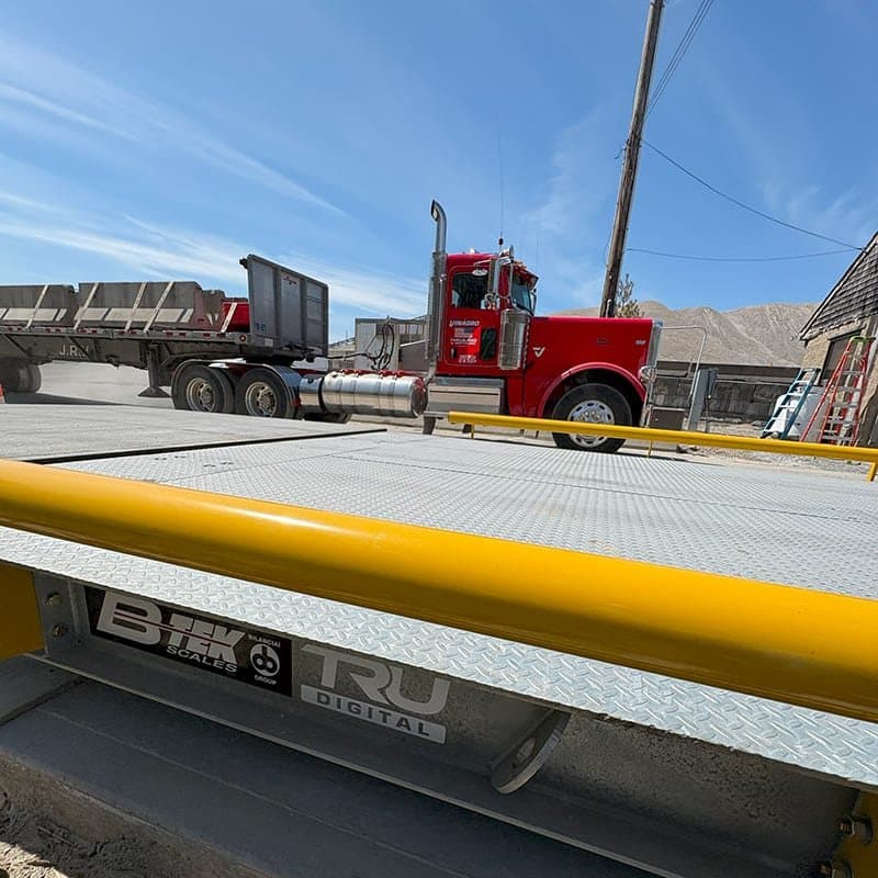 Truck scale deck with a digital load cell nameplate on the side rail, loaded truck waiting behind